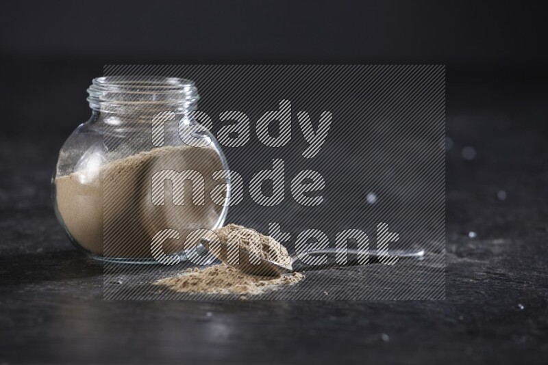 A glass spice jar full of garlic powder with a filled metal spoon on a textured black flooring