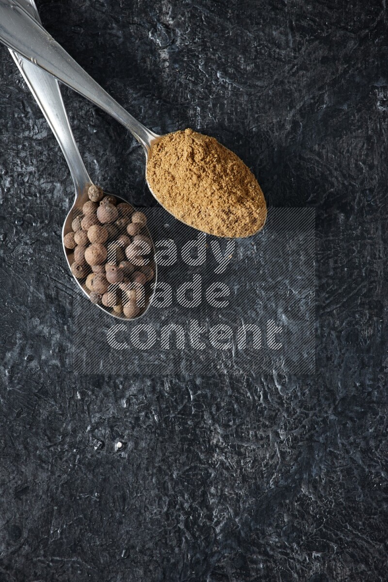 2 metal spoons full of allspice powder and whole balls on a textured black flooring
