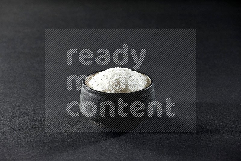 A black pottery bowl full of desiccated coconut on a black background in different angles