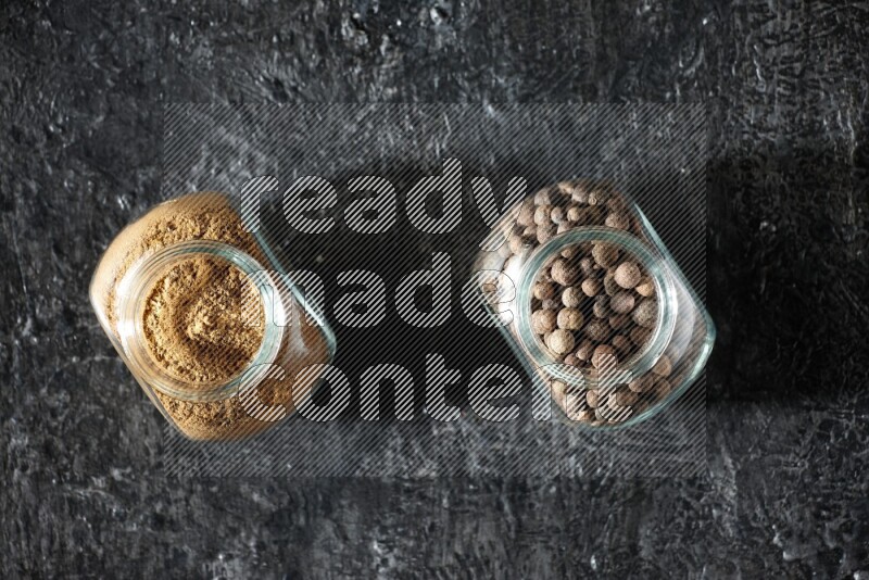 2 glass spice jars full of allspice powder and whole balls on a textured black flooring