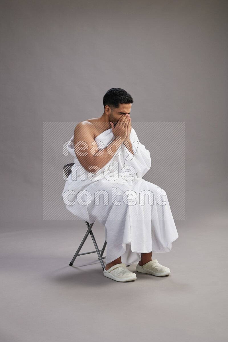 A man wearing Ehram sitting on chair showing how to sneeze on gray background
