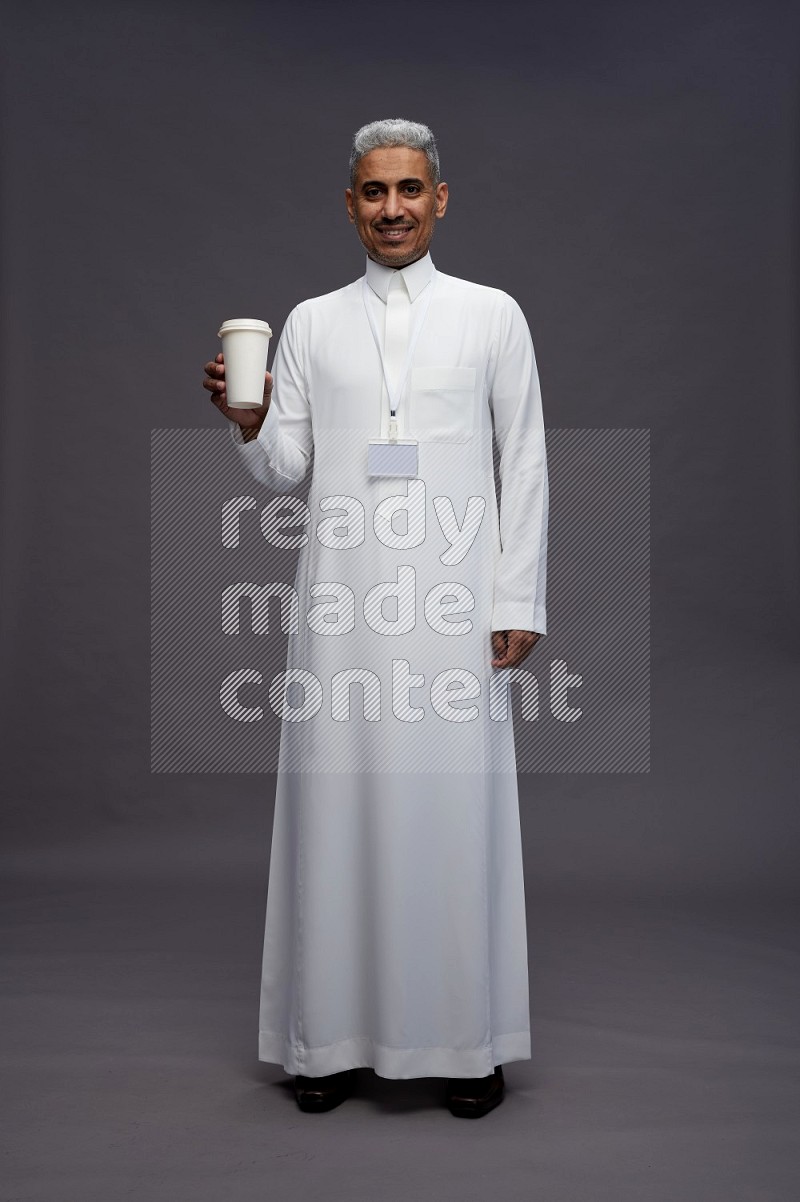 Saudi man wearing thob with neck strap employee badge standing holding paper cup on gray background