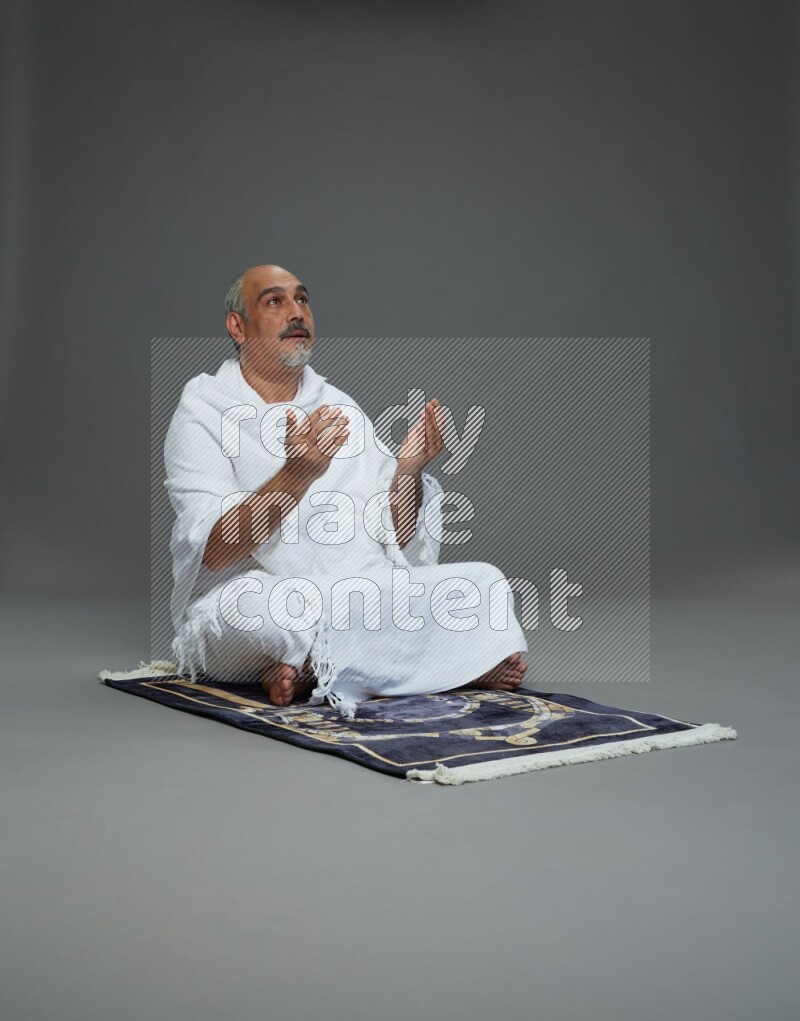 A man wearing Ehram sitting on prayer mat dua'a on gray background