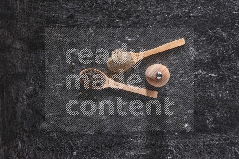 Black pepper powder and black pepper beads in wooden spoons and wooden grinder on a textured black flooring