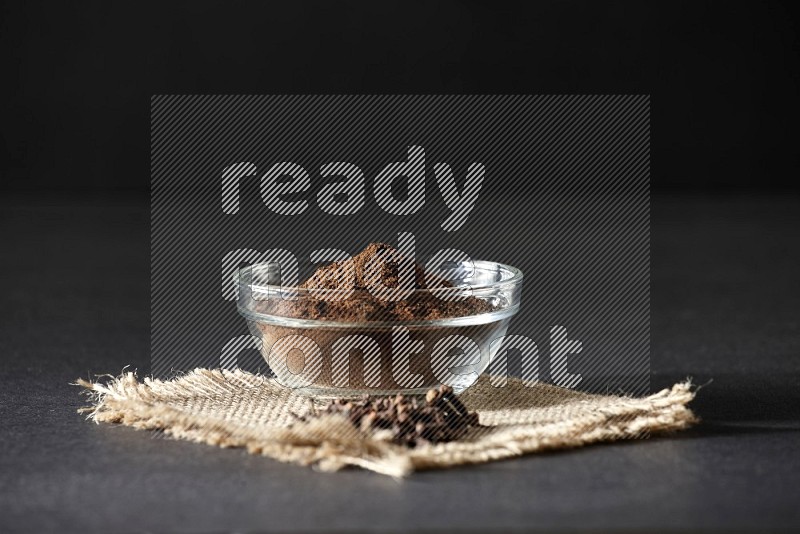 A glass bowl full of cloves powder with cloves grains on a burlap piece on a black flooring