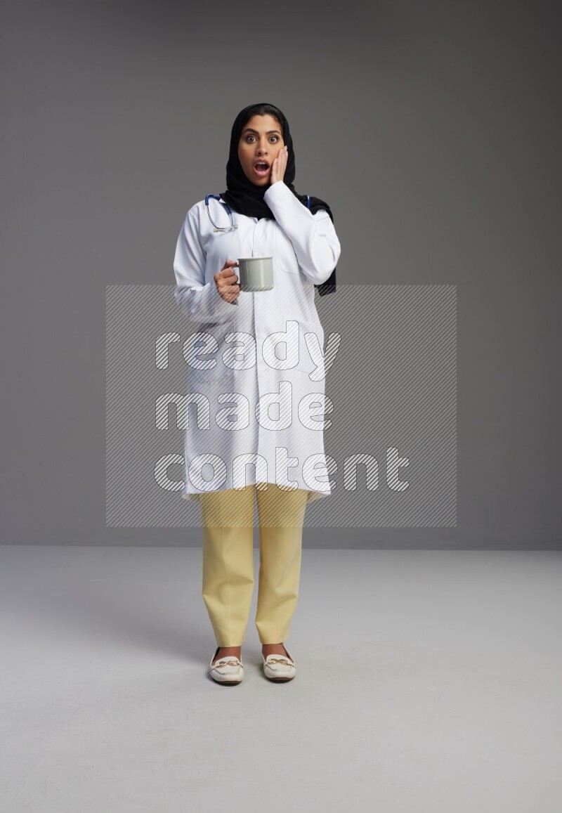 Saudi woman wearing lab coat with stethoscope standing holding mug on Gray background