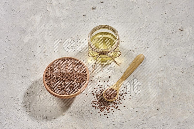 A wooden bowl and spoon full of flax seeds and a glass jar of flaxseeds oil on a textured white flooring