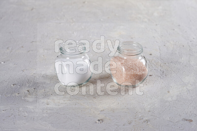 2 glass jars one is full of fine himalayan salt and the other with table salt on white background
