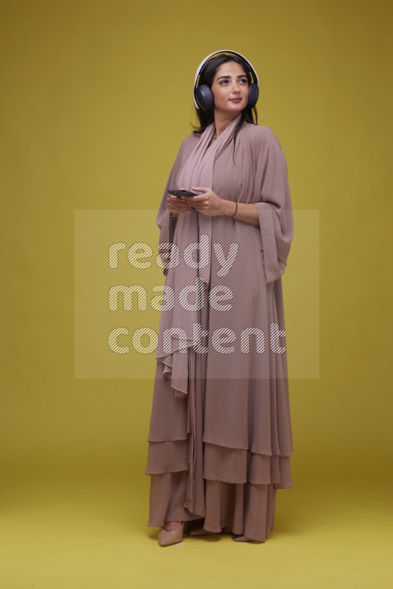 A Saudi woman Listing to Music on a Yellow Background wearing Brown Abaya