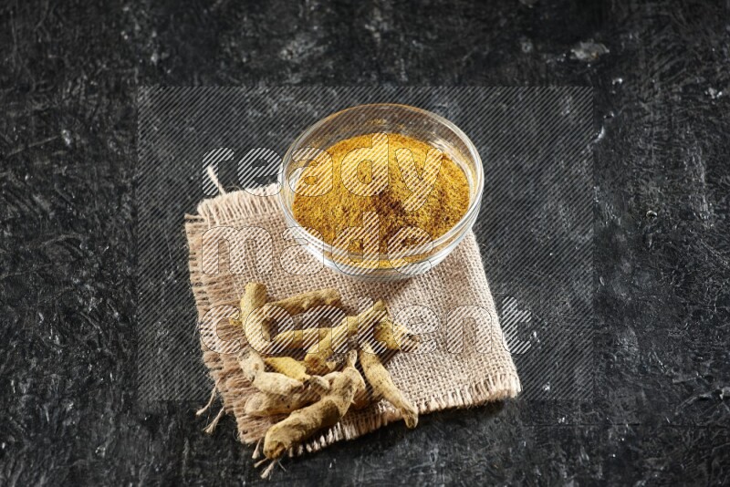 A glass bowl full of turmeric powder with dried turmeric fingers on a burlap fabric on textured black flooring
