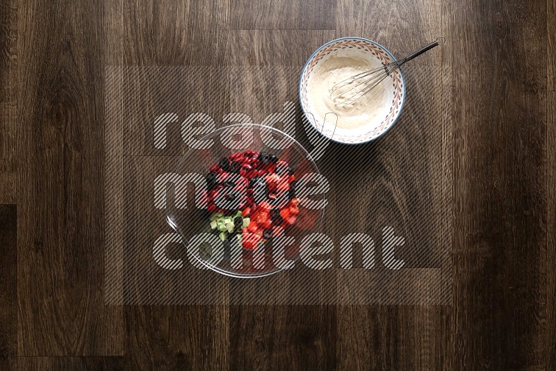 A bowl full of salad (avocado, tomatoes, red beans, olives, bell pepper, corn, lettuce) and bowl of salad dressing on wooden background