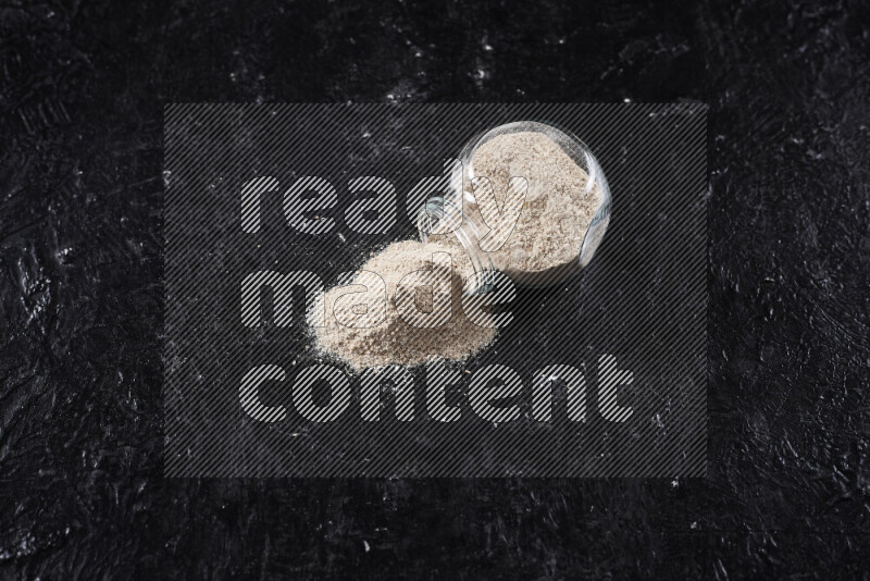 A glass jar full of onion powder flipped with some spilling powder on black background