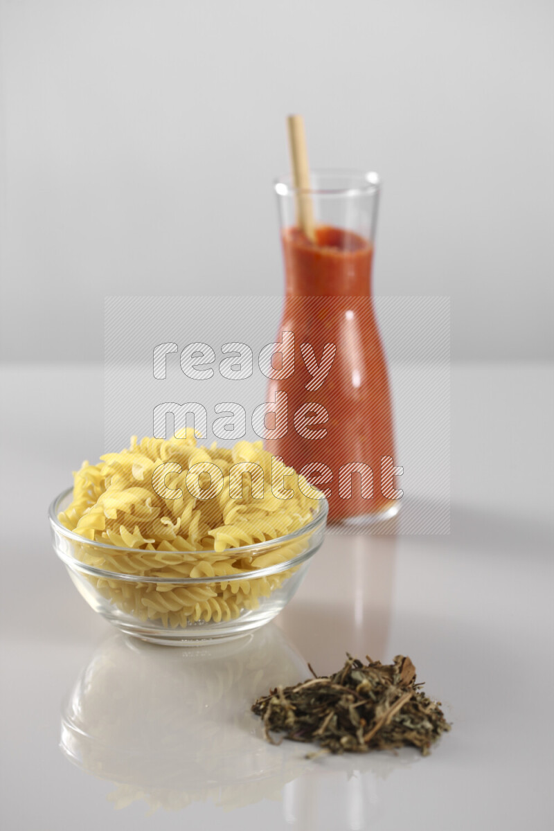 Raw pasta with tomatoe pasta with different ingredients such as cherry tomatoes, basil, garlic, bay laurel, cardamom, white pepper, black pepper, red chilis and wheat stalks on light grey background