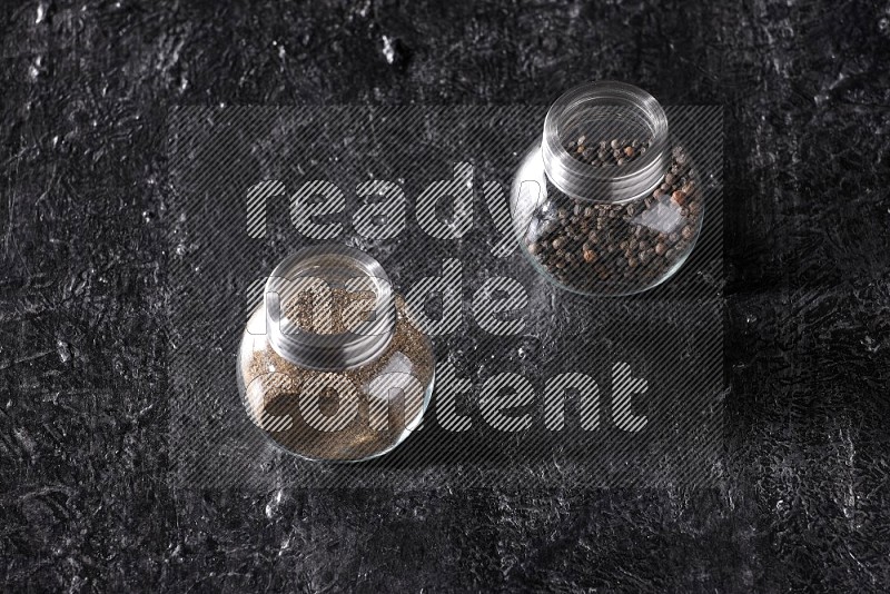2 glass spice jars full of black pepper powder and black pepper beads on textured black flooring