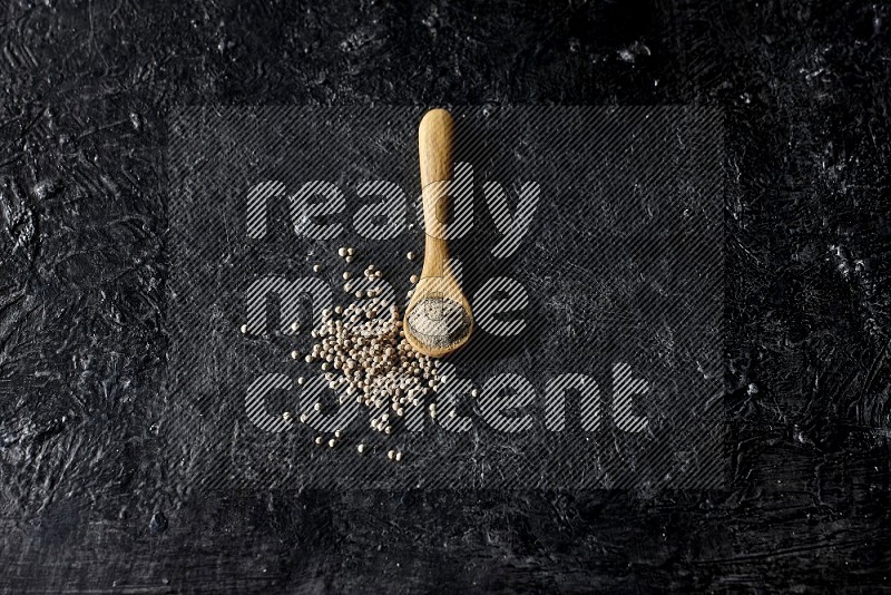 A wooden spoon full of white pepper powder with white pepper beads on textured black flooring