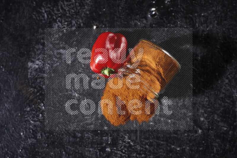 A glass jar full of ground paprika powder flipped with some spilling powder on black background