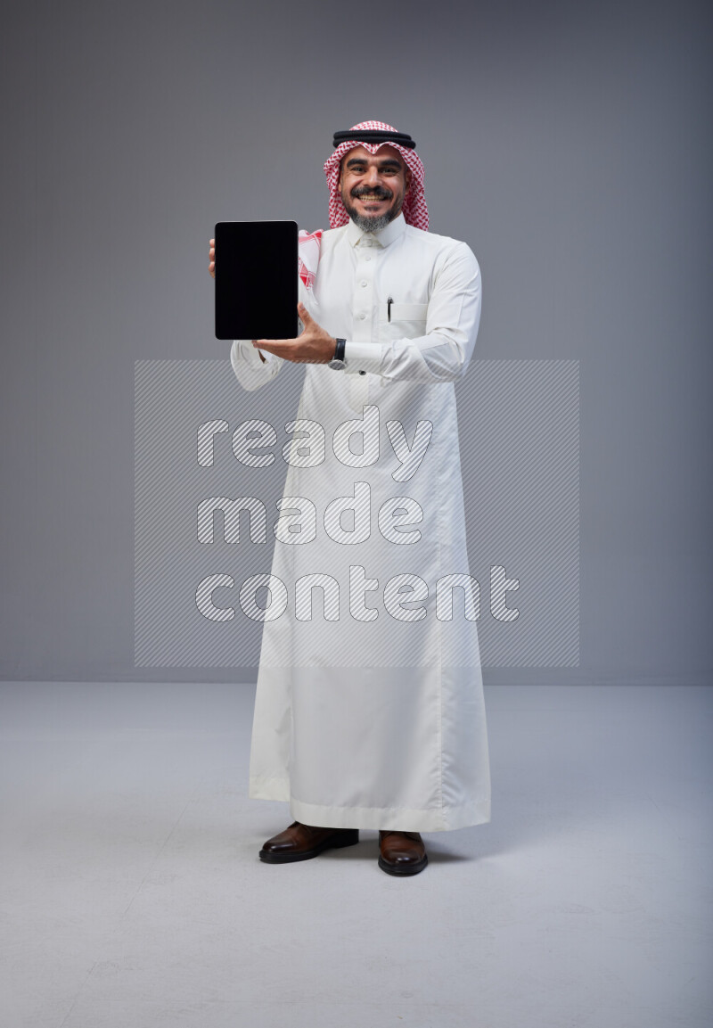 Saudi man Wearing Thob and red Shomag standing showing tablet to camera on Gray background