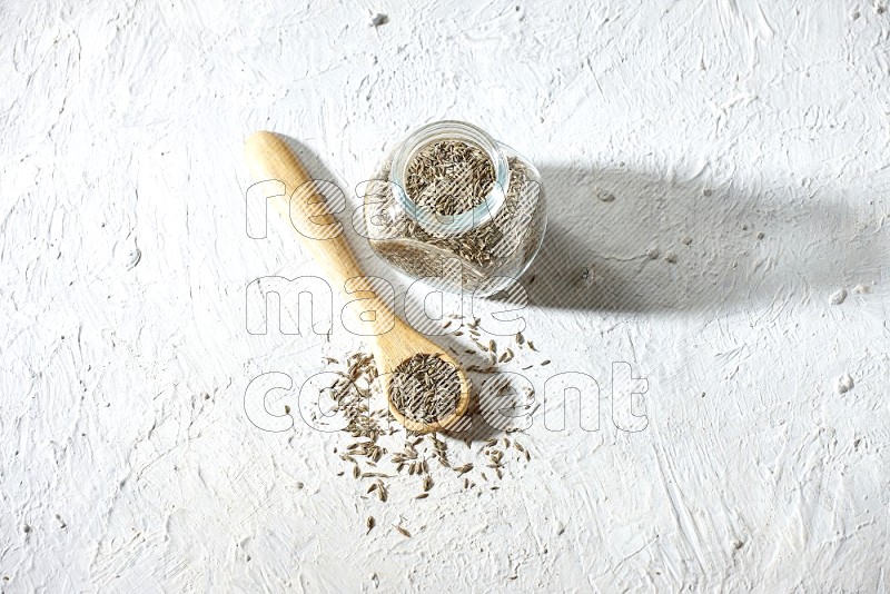 A glass spice jar and wooden spoon full of cumin seeds on textured white flooring