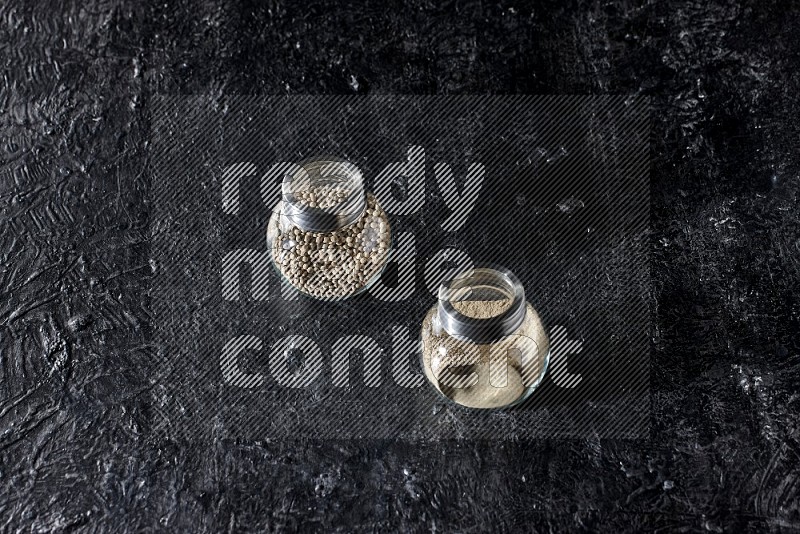 2 Herbal glass jars full of white pepper beads and powder on textured black flooring