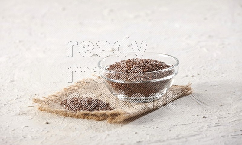 A glass bowl full of flax seeds and a bunch of seeds on burlap fabric on a textured white flooring