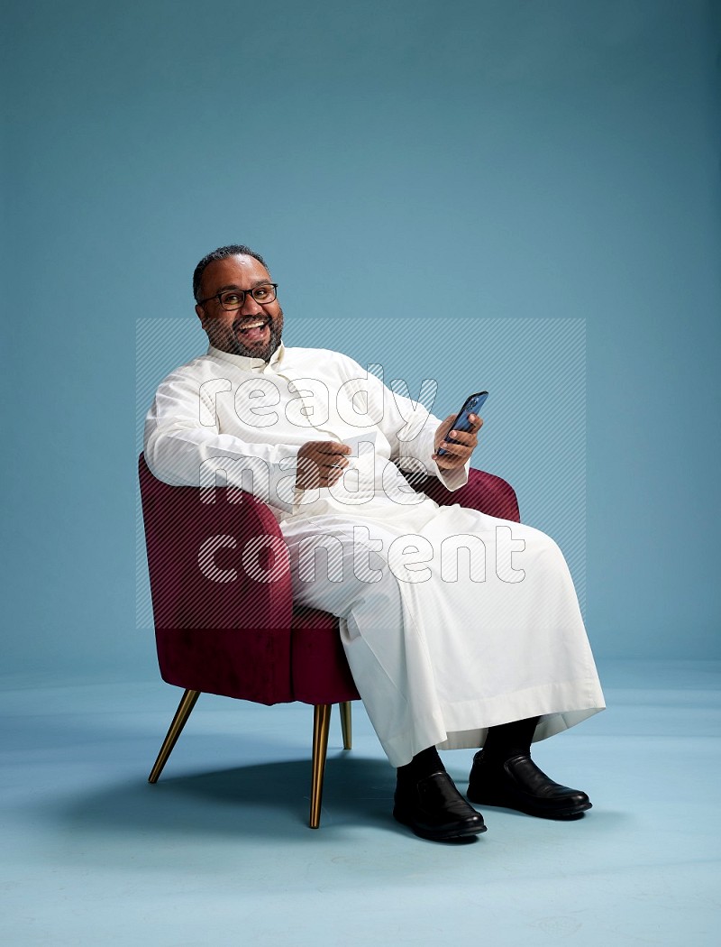 Saudi Man without shimag sitting on chair holding ATM while talking on phone on blue background