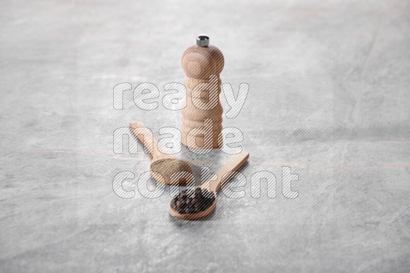 2 wooden spoons, one full of black pepper powder and the other full of black pepper beads and wooden grinder on a marble flooring