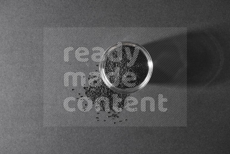 A glass bowl full of black seeds and seeds spreaded beside it on a black flooring