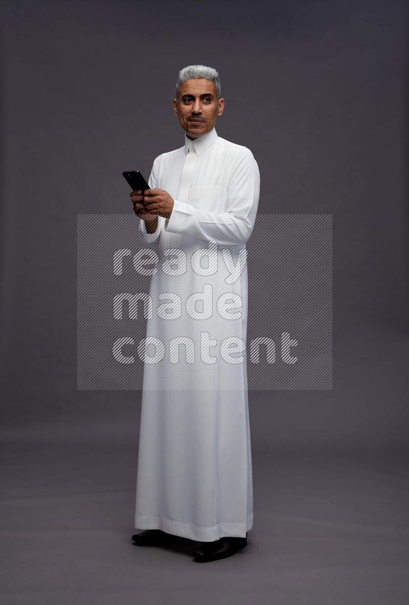 Saudi man wearing thob standing texting on phone on gray background