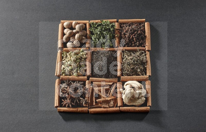 9 مربعات من أعواد القرفة ممتلئة بالشاي في المنتصف محاطة بجوزة الطيب والنعناع المجفف والقرنفل والريحان المجفف والزنجبيل المجفف والقرفة واليانسون والهيل على أرضية سوداء