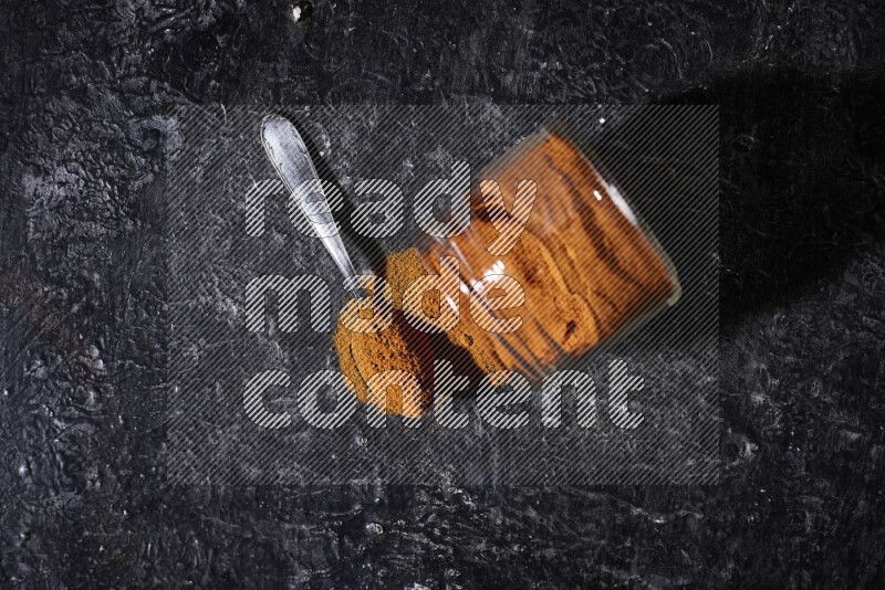 A glass jar full of ground paprika powder flipped with some spilling powder on black background