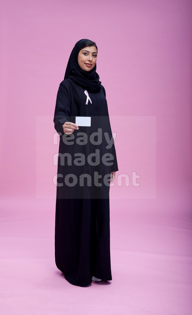 Saudi woman wearing pink ribbon on Abaya standing holding ATM card on pink background