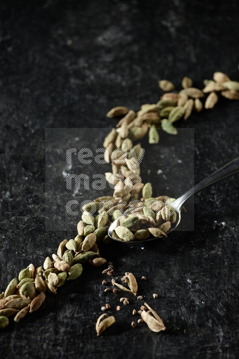 A Metal spoon full of cardamom seeds and some seeds beside it on a textured black flooring