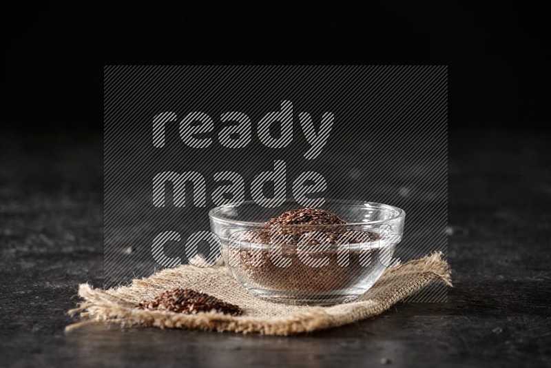 وعاء زجاجي ممتلئ بحبوب بذر الكتان مع كومة من البذور على قطعة من القماش على أرضية سوداء