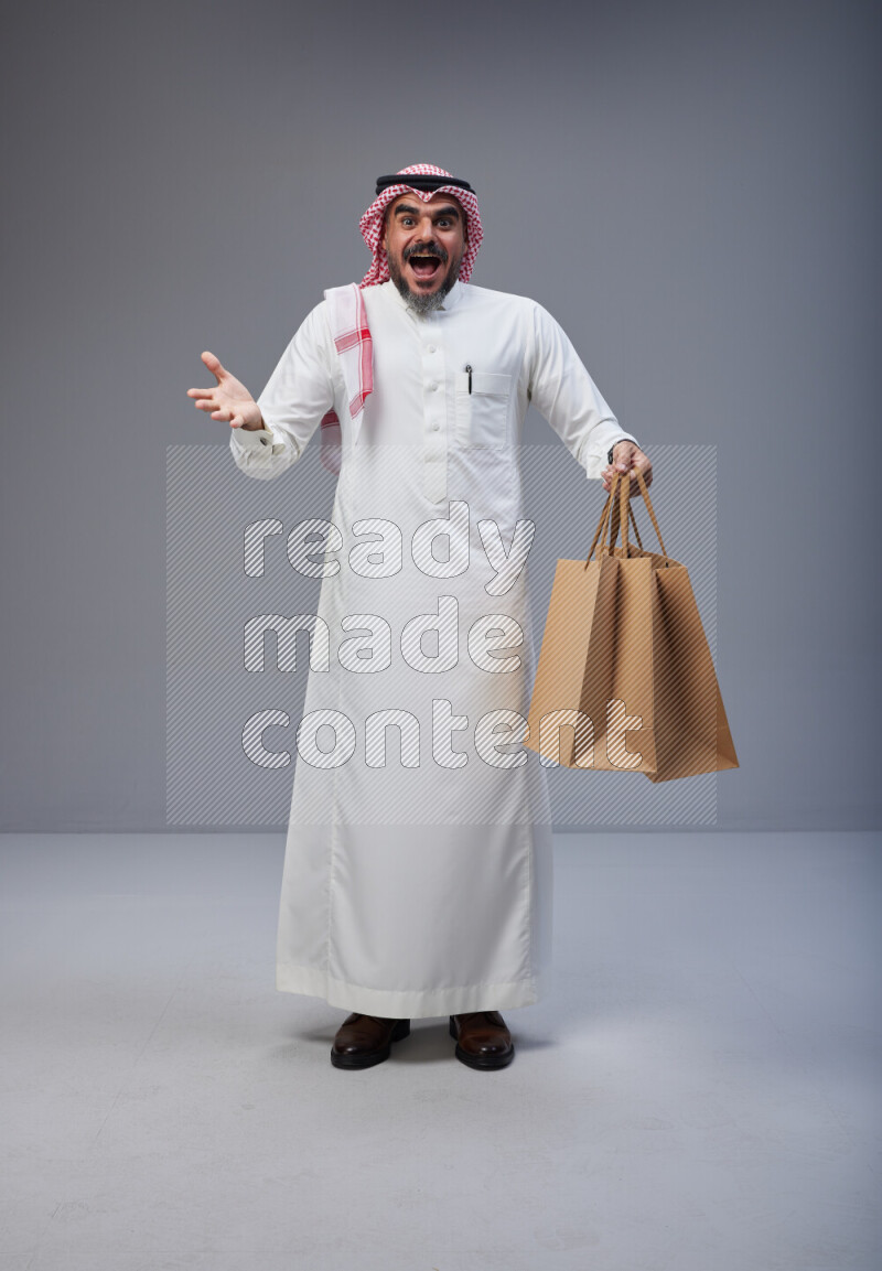Saudi man Wearing Thob and red Shomag standing holding shopping bag on Gray background