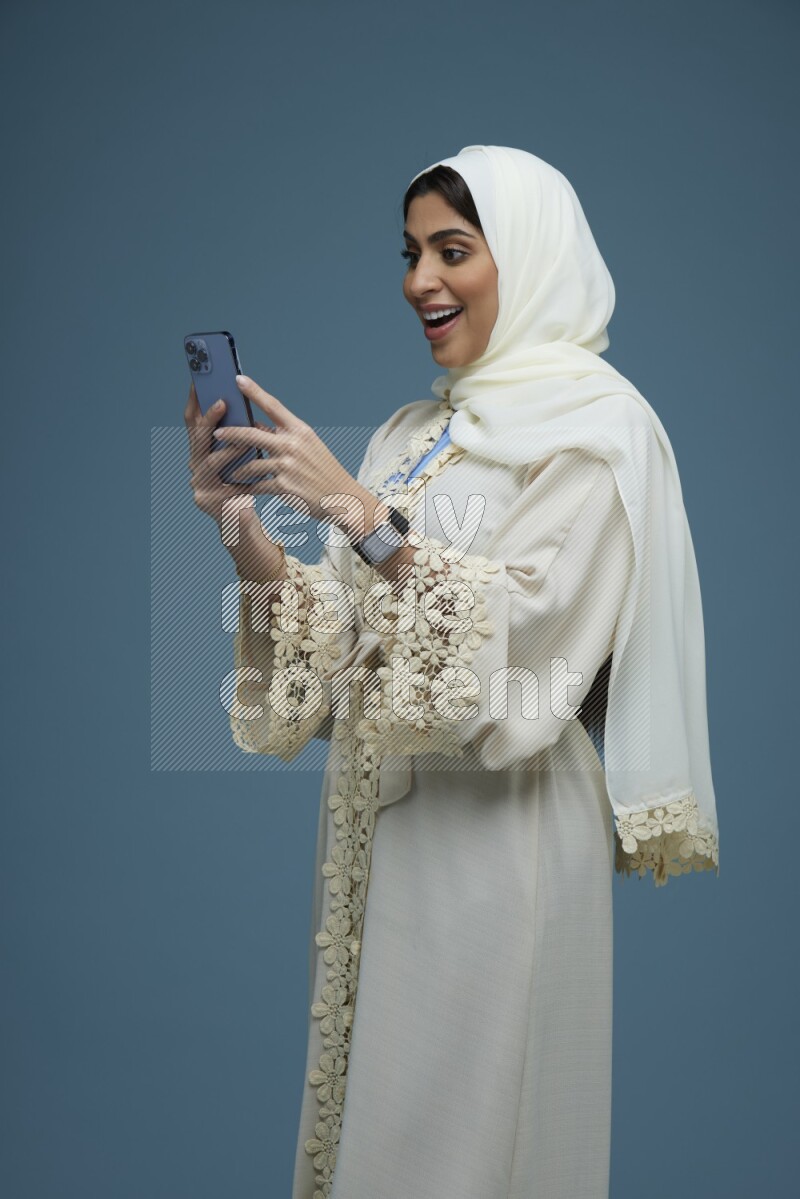 A Saudi woman Texting in a blue background wearing an off-white Abaya Hijab