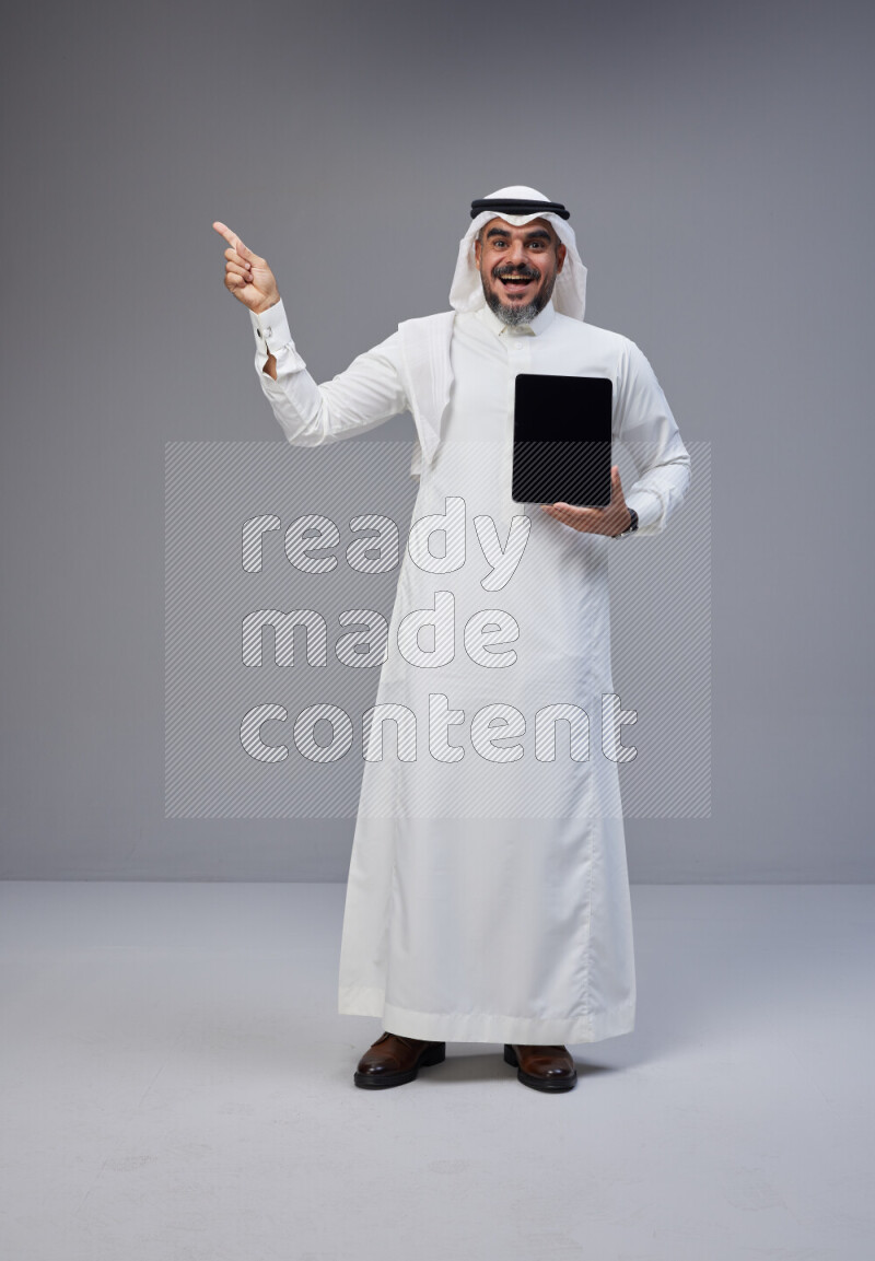 Saudi man Wearing Thob and white Shomag standing showing tablet to camera on Gray background