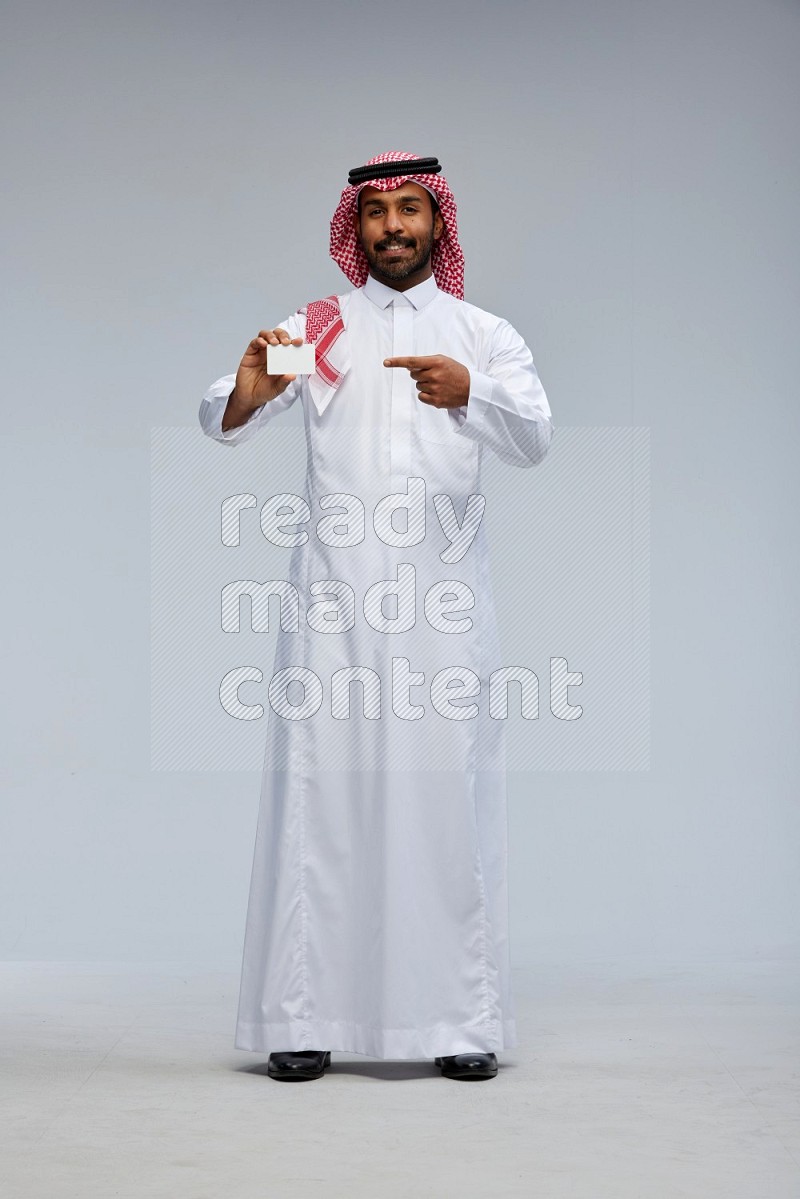 Saudi man wearing thob and shomag standing holding ATM card on gray background