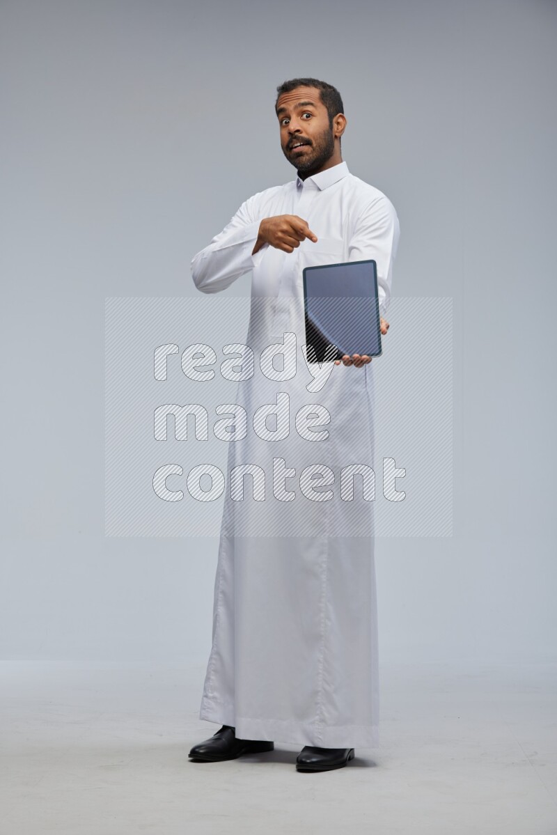 Saudi man wearing Thob standing showing tablet to camera on Gray background