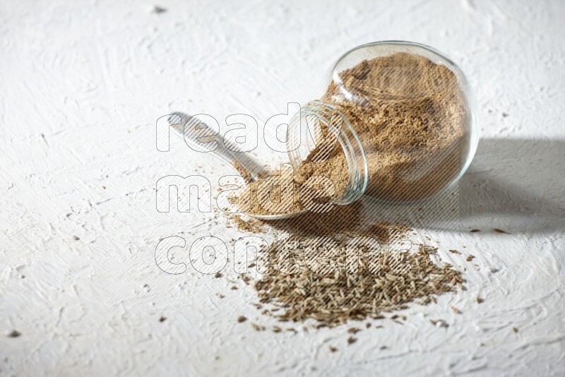 A glass spice jar and metal spoon full of cumin powder and the jar flipped and powder spilled out with cumin seeds on textured white flooring