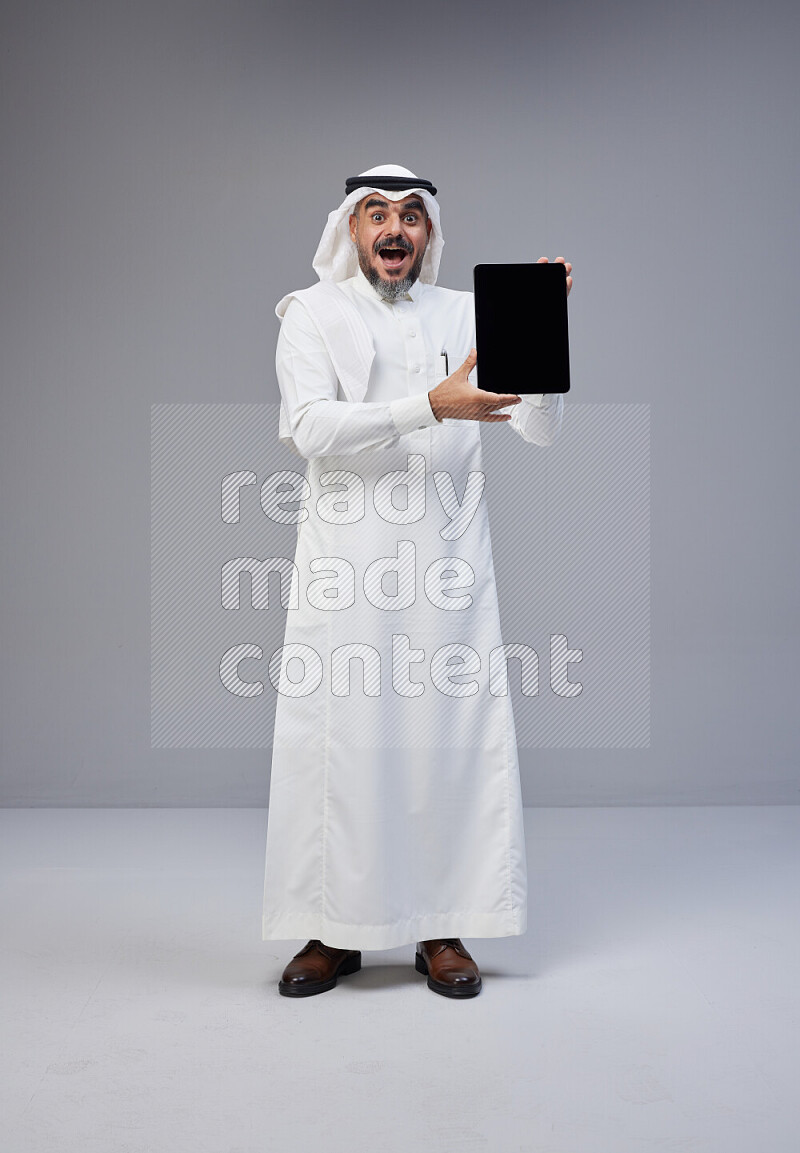 Saudi man Wearing Thob and white Shomag standing showing tablet to camera on Gray background