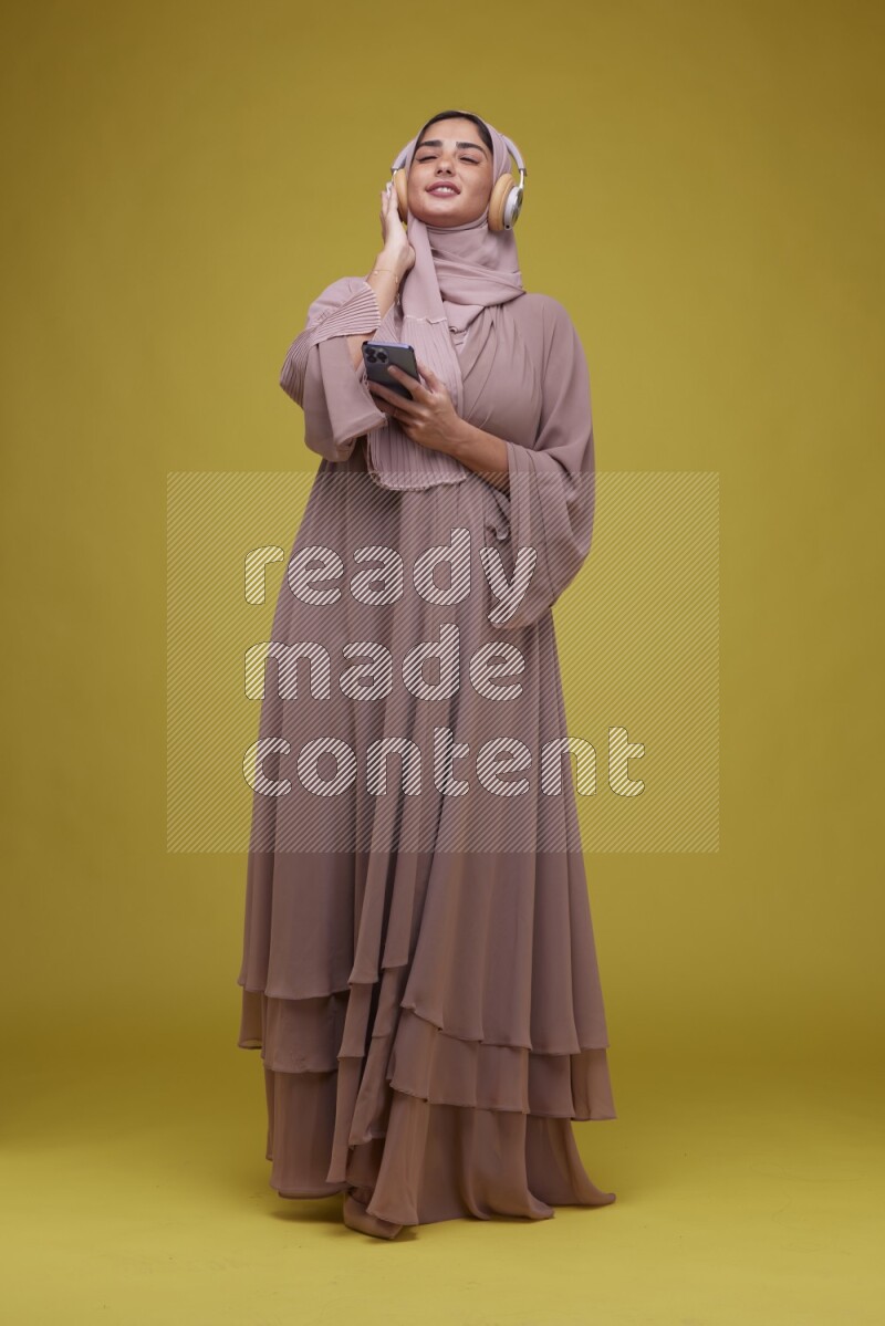 A woman Listening to Music on a Yellow Background wearing Brown Abaya with Hijab