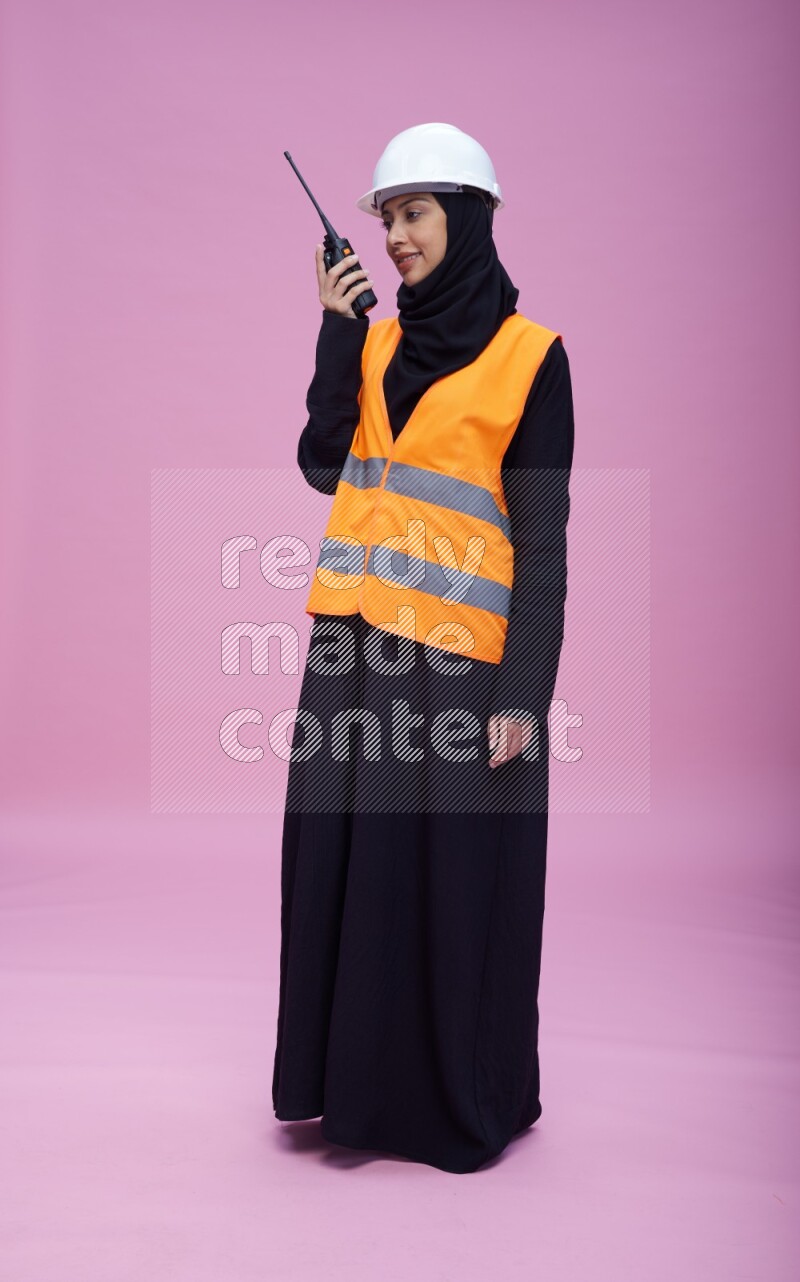 Saudi woman wearing Abaya with engineer vest and helmet standing holding walkie-talkie on pink background
