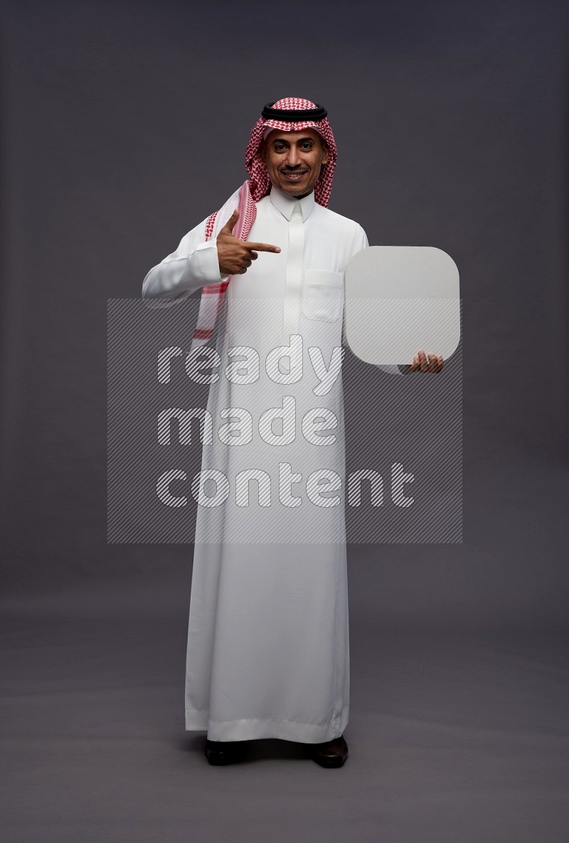 Saudi man wearing thob and shomag standing holding social media sign on gray background