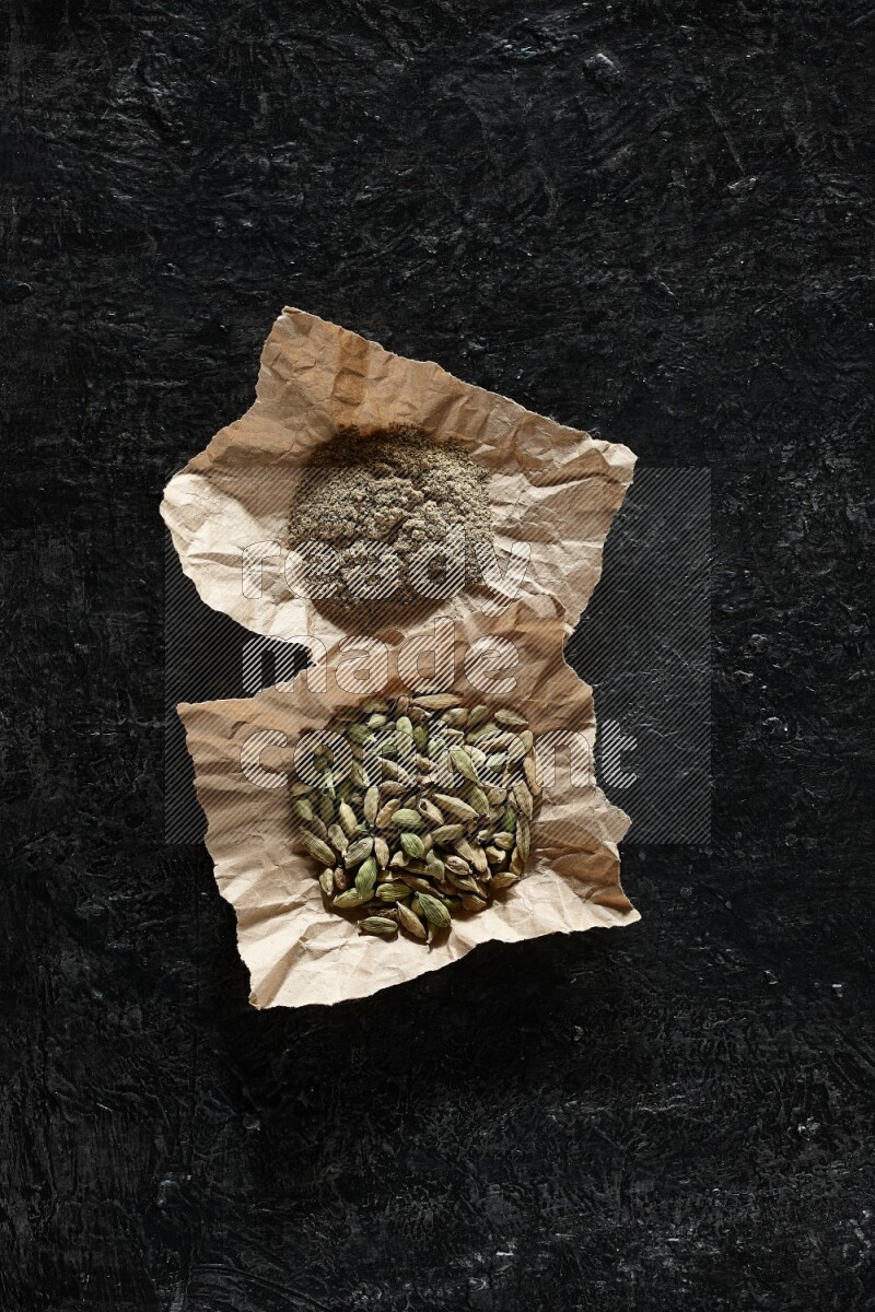 Cardamom seeds and cardamom powder in 2 crumpled pieces of paper on textured black flooring