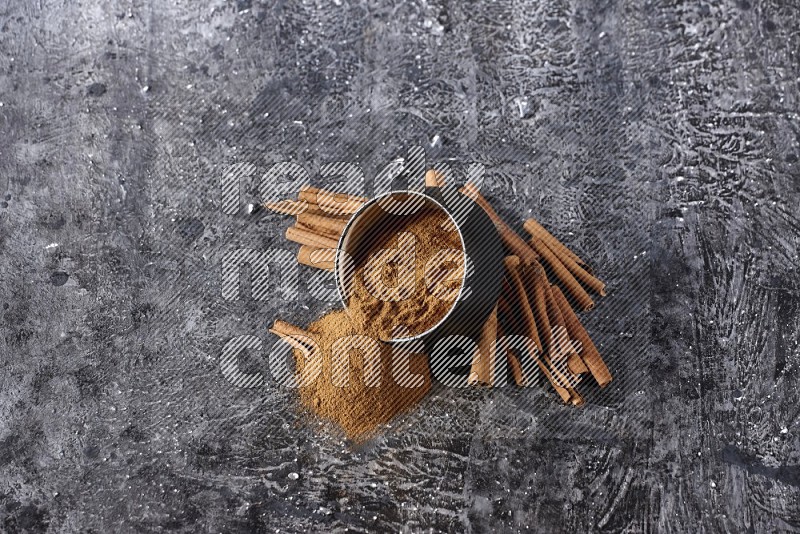 Black pottery bowl over filled with cinnamon powder and cinnamon sticks around the bowl on a textured black background