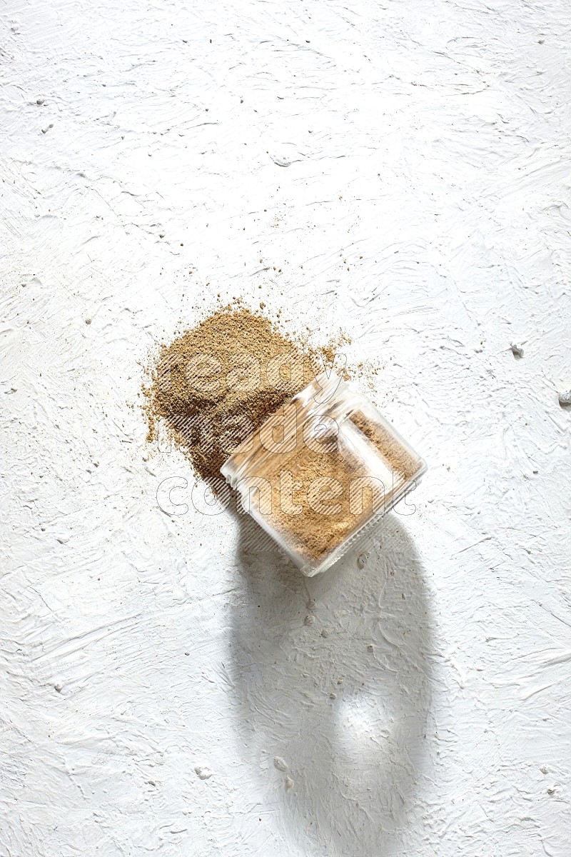 A flipped glass jar full of cumin powder and powder spilled out on textured white flooring