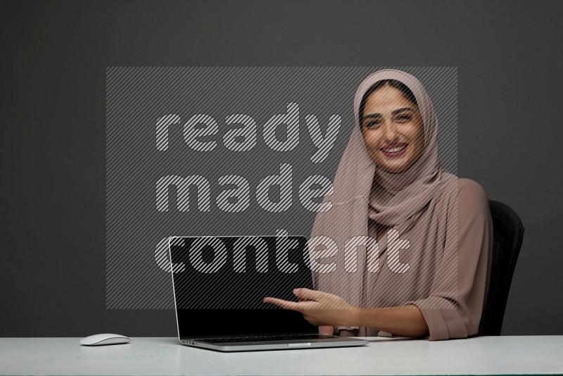 A woman Sitting on her desk  Pointing at her laptop on a Gray Background wearing Brown Abaya with Hijab