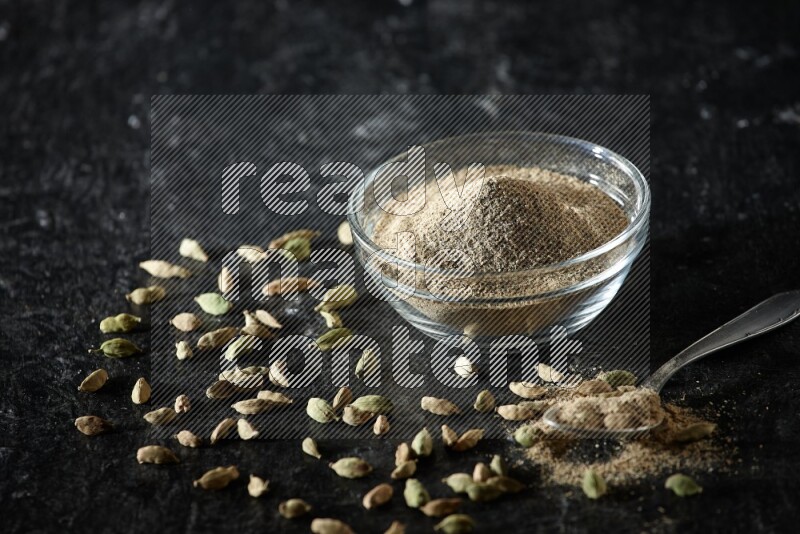 A glass bowl and a metal spoon full of cardamom powder with cardamom seeds beside them on textured black flooring