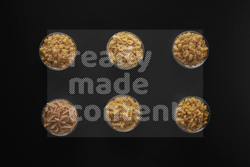 Different pasta types in 6 glass bowls on black background