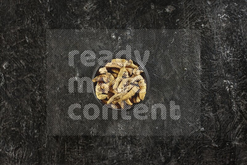 A black pottery bowl full of dried turmeric whole fingers on textured black flooring